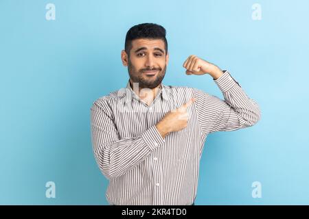 Uomo d'affari forte bearded alzando le mani che mostrano il potere, indicando i suoi bicipiti, ritenendo indipendente forte con sguardo fiero, portando la camicia a strisce. Studio in interni isolato su sfondo blu. Foto Stock