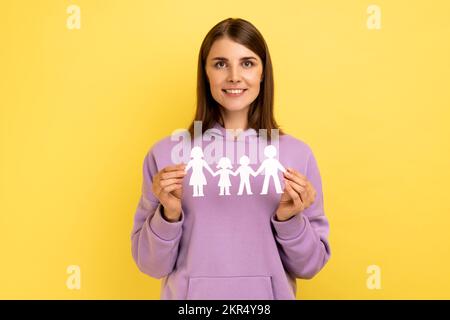 Genere sorridente donna positiva che tiene la gente della catena della carta che tiene nelle mani, famiglia felice, rapporti, paternità, con la felpa con cappuccio viola. Studio al coperto isolato su sfondo giallo. Foto Stock