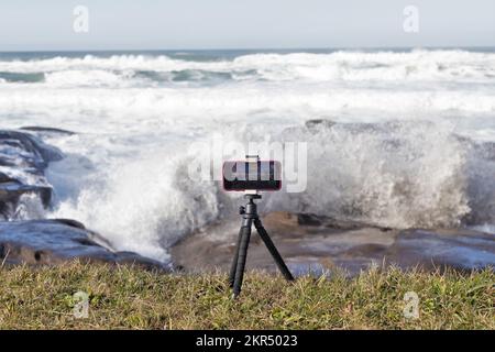 Un iPhone su un piccolo treppiede cattura riprese di onde enormi durante una marea reale sulla costa rocciosa a Yachats, Oregon. Foto Stock