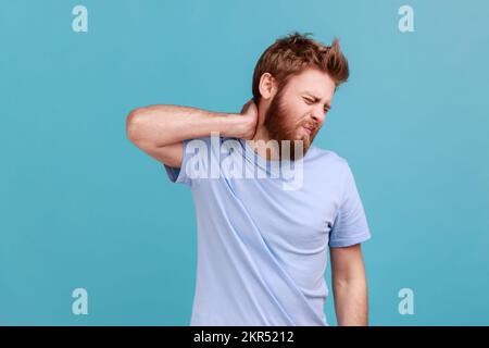 Ritratto di uomo insano esausto malsano che massaggia il collo dolorante con espressione di sofferenza insopportabile, con problemi alla colonna vertebrale, mal di schiena. Studio in interni isolato su sfondo blu. Foto Stock