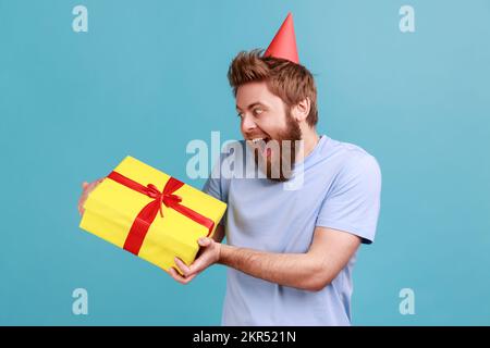 Ritratto di uomo sopportato estremamente felice essendo molto felice di ottenere il presente da un amico o da una ragazza, tenendo la scatola del regalo, esprimendo l'eccitamento. Studio in interni isolato su sfondo blu. Foto Stock