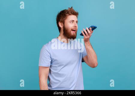 Ritratto di un uomo ottimista che registra un messaggio vocale o parla con un assistente online sul suo smartphone tenendo il telefono vicino alla bocca. Studio in interni isolato su sfondo blu. Foto Stock