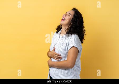 Ritratto di donna overjoyed con i capelli ondulati scuri che ridono felicemente a qualcosa mantiene le mani sul ventre, sorride ampiamente, esprimendo emozioni positive.Studio interno girato isolato su sfondo giallo. Foto Stock