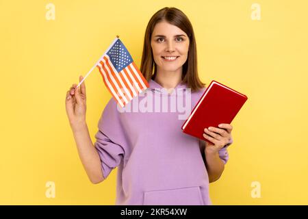 Ritratto di attraente studente donna con capelli scuri in possesso di libro e bandiera americana, istruzione all'estero negli Stati Uniti, con cappuccio viola. Studio al coperto isolato su sfondo giallo. Foto Stock