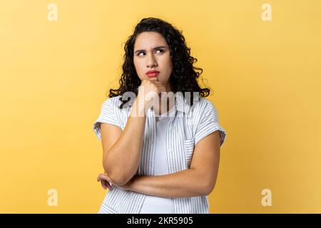 HMM, ho bisogno di pensare. Ritratto di donna con capelli ondulati scuri che tengono il mento e la musa, guardando via con premurosa espressione facciale. Studio al coperto isolato su sfondo giallo. Foto Stock