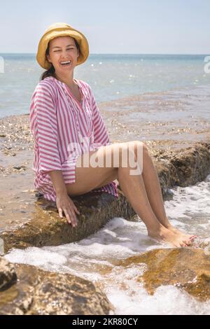Una giovane donna gioiosa che ride seduto su una spiaggia di pietra circondata da spruzzi d'acqua onde schiumose Foto Stock