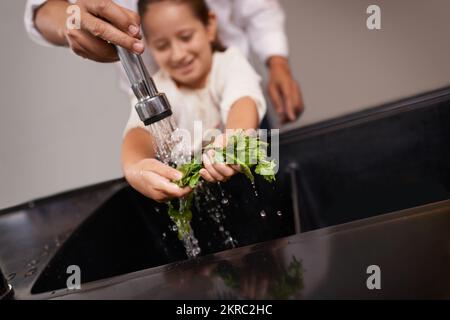 Dandole una mano d'aiuto. una bambina che lava le verdure al lavandino con l'aiuto di suo nonno. Foto Stock