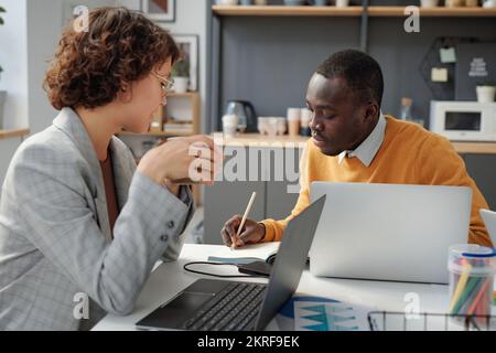 Giovane donna d'affari che beve caffè mentre si siede al tavolo con la sua collega che lavora sui computer in ufficio Foto Stock