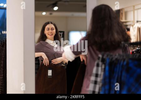 Shopping in seconda mano. Felice sorridente giovane donna caucasica provare vestiti e guarda lo specchio. Concetto di acquisto e vendita. Foto Stock