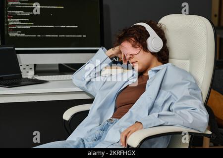 Giovane donna d'affari in maschera rilassarsi sulla sedia sul suo posto di lavoro mentre si ascolta la musica in cuffie Foto Stock