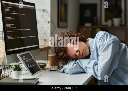 Sviluppatore IT in maschera che dorme sul suo posto di lavoro dopo un duro lavoro con il software sul computer Foto Stock