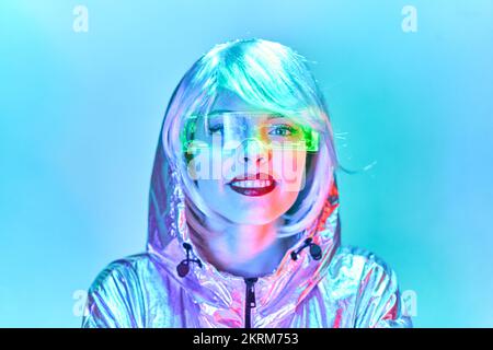 Giovane donna sorridente in parrucca con occhiali intelligenti per realtà aumentata con schermo virtuale che visualizza le icone mentre si è in piedi in studio con blu al neon brillante Foto Stock