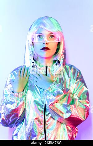 Giovane donna in parrucca con occhiali intelligenti per realtà aumentata con schermo virtuale che visualizza icone mentre si è in piedi in studio con luci blu al neon incandescenti Foto Stock