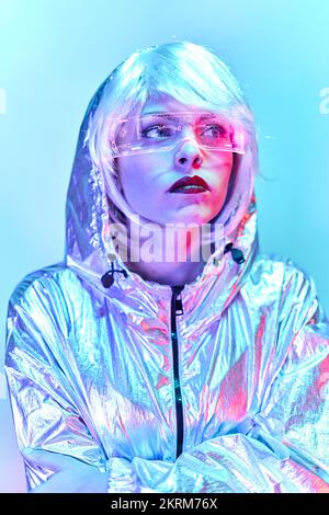 Giovane donna in parrucca con occhiali intelligenti per realtà aumentata con schermo virtuale che visualizza icone mentre si è in piedi in studio con luci blu al neon incandescenti Foto Stock