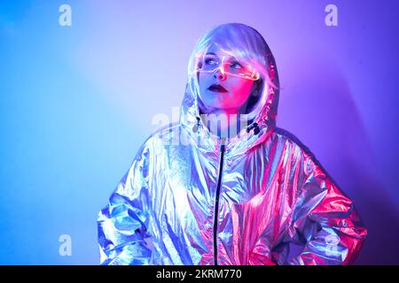 Giovane donna in parrucca con occhiali intelligenti per realtà aumentata con schermo virtuale che visualizza icone mentre si è in piedi in studio con luci blu al neon incandescenti Foto Stock
