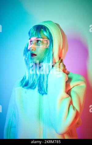 Giovane donna in parrucca con occhiali intelligenti per realtà aumentata con schermo virtuale che visualizza icone mentre si è in piedi in studio con luci blu al neon incandescenti Foto Stock