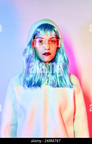 Giovane donna in parrucca con occhiali intelligenti per realtà aumentata con schermo virtuale che visualizza icone mentre si è in piedi in studio con luci blu al neon incandescenti Foto Stock