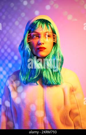 Giovane donna in parrucca con occhiali intelligenti per realtà aumentata con schermo virtuale che visualizza icone mentre si è in piedi in studio con luci rosa al neon scintillanti Foto Stock