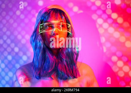 Giovane donna in parrucca con occhiali intelligenti per realtà aumentata con schermo virtuale che visualizza icone mentre si è in piedi in studio con luci rosa al neon scintillanti Foto Stock