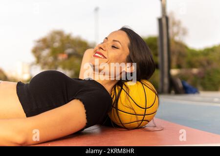 Vista laterale di una donna sorridente in cima raccolto sdraiata a terra sul campo con basket sotto la testa mentre riposa dopo la partita Foto Stock
