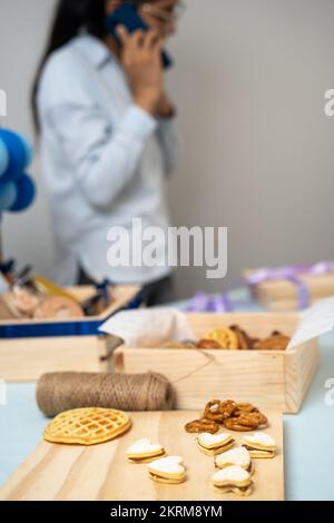 Crop femmina che ha conversazione sul cellulare, mentre la preparazione dolce presente con cialde pretzel e biscotti in scatole Foto Stock