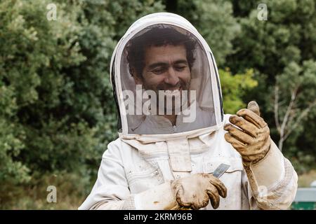 Sorridente Beekeeper maschio felice in uniforme protettiva bianca e guanti tenendo raschiatore utensile apertura alveare verde con api mentre si lavora in apiario su su Foto Stock