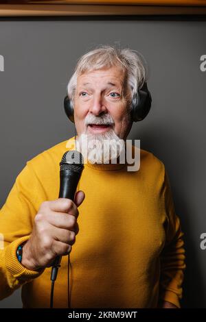 Allegro uomo anziano bearded in giallo maglione cantando in microfono su sfondo grigio Foto Stock