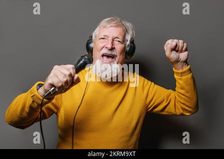 Allegro uomo anziano bearded in giallo maglione clenching pugno mentre cantano in microfono contro sfondo grigio Foto Stock