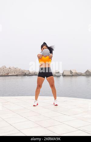 Corpo pieno di determinata giovane femmina con pigtail in abbigliamento sportivo facendo esercizio con kettlebell vicino al mare Foto Stock
