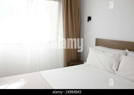 Comodo letto coperto da lenzuola bianche collocato in una moderna camera d'albergo minimalista nelle giornate di sole Foto Stock