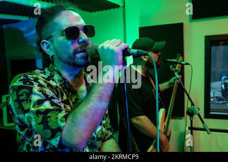 Uomo che suona la chitarra elettrica mentre il cantante canta la canzone nel microfono durante le prove in studio scuro con illuminazione al neon brillante Foto Stock
