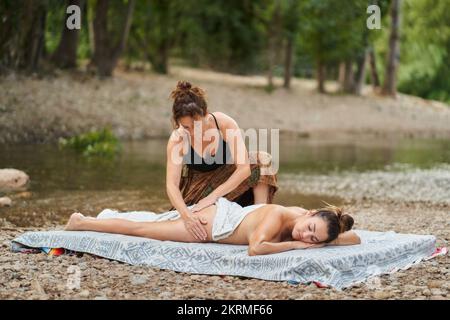 Massaggiatrice adulta in top nero facendo massaggio natiche di cliente femminile sdraiato con gli occhi chiusi sul tappetino vicino allo stagno Foto Stock