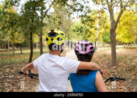 Vista posteriore ritagliata degli adolescenti in abiti casual e caschi riposati nel parco autunnale in piedi sul sentiero durante la pausa dalla guida Foto Stock