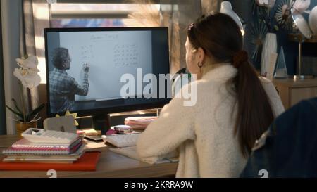 Giovane ragazza asiatica si siede al tavolo a casa accogliente e scrive compiti a casa. Schoolgirl ascolta la lezione online di un insegnante maturo e studia sul computer. Concetto di apprendimento a distanza. Studio remoto. Vista posteriore. Foto Stock