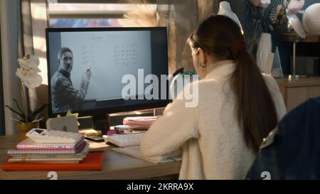 Giovane ragazza asiatica si siede al tavolo a casa accogliente e scrive compiti a casa. Schoolgirl ascolta la lezione online di un insegnante maturo e studia sul computer. Concetto di apprendimento a distanza. Studio remoto. Vista posteriore. Foto Stock