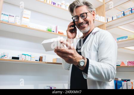 Il farmacista legge l'etichetta di un farmaco mentre parla con un paziente al telefono. Operatore sanitario senior che conferma una prescrizione tramite una telefonata. M Foto Stock