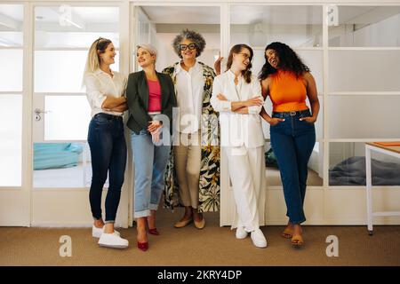 Gruppo di donne d'affari di successo sorridendo felicemente mentre si levano in piedi insieme in un ufficio creativo. Donne d'affari multiculturali che lavorano come una squadra in un al Foto Stock