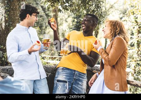 gruppo multirazziale di amici ridendo e divertendosi alla festa, gruppo misto di fascia d'età che celebra qualcosa mangiando spuntino e bevendo birre Foto Stock