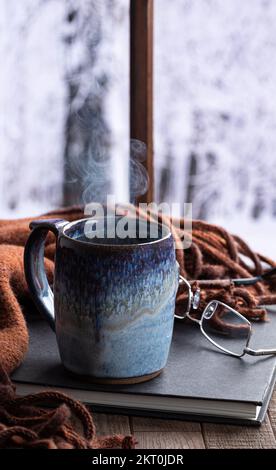 Una tazza calda di caffè o tè fumante vicino a una finestra con sfondo invernale innevato Foto Stock