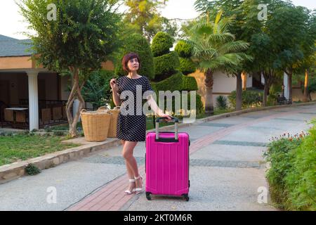 Una donna con una grande valigia rosa brillante è in piedi sulla strada non sapendo dove andare dopo. Foto Stock