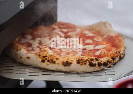 Cucinare o cuocere la pizza Margherita calda con salsa di pomodoro e mozzarella con una pala in un forno elettrico o a gas, con fumo, primo piano Foto Stock