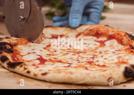 Mano con guanto di plastica tagliando pizza Margherita calda con salsa di pomodoro e mozzarella con taglierina su un asse di legno, primo piano Foto Stock