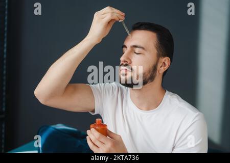 Uomo caucasico bearded applicando il siero di acne di bellezza, seduto al mattino nel soggiorno. Terapia nutriente, concetto di cura della pelle. Foto Stock