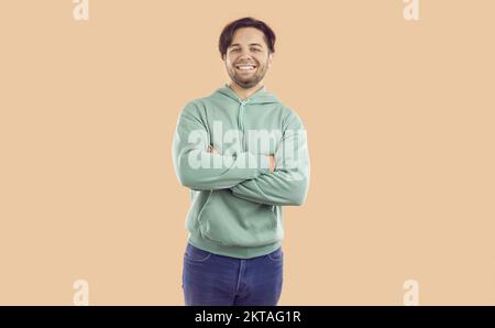 Ritratto di felice uomo brunetto sorridente in felpa cappuccio menta e jeans su sfondo beige guardando la macchina fotografica. Foto Stock