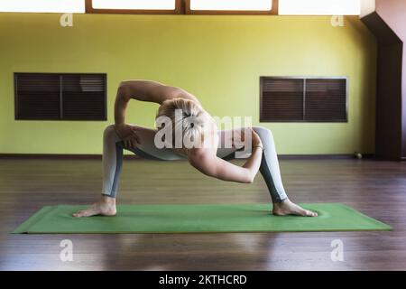 Donna che pratica yoga, facendo torsione in rudrasana, allenandosi mentre si trova in piedi su un tappetino in studio Foto Stock