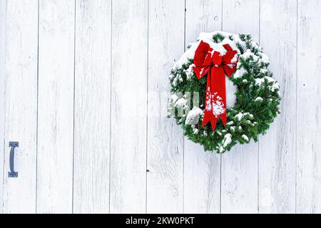 Neve spolverata ago di pino wreath Natale e arco rosso sulla grande tavola intemperie fienile porta. Rustico, naturale, tradizionale paese Natale. Copia spazio. Foto Stock