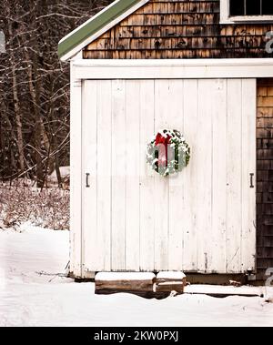 Corona d'ago di pino innevata e arco rosso sulla porta di legno intaccato e imbiancato del piccolo fienile. Rustico, rurale, sottomesso, tradizionale paese Natale. Foto Stock