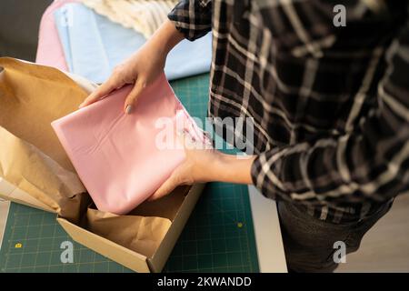 Mani di giovane donna venditore di sartoria del negozio in linea che mette il panno piegato nella scatola prima dell'imballaggio e dell'invio al cliente. Piccolo proprietario in linea di affari Foto Stock