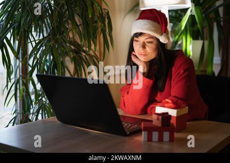 Donna triste hipster in cappello di Santa seduta a casa a Natale. Dura giornata. Festa virtuale della casa di Natale. Spazio di copia Foto Stock