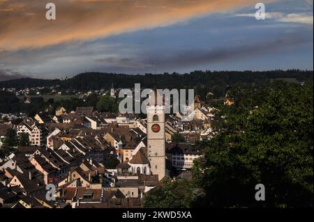 Cielo arancione e blu tempestoso sopra le tane, i tetti in tegole e la torre gotica dell'orologio Kirche St Johann nella Altstadt o nella città vecchia di Schaffhausen, nel nord della Svizzera. Le strade prive di traffico sono fiancheggiate da molte belle gilde gotiche, rinascimentali e barocche o case mercantili con finestre a bovindo e facciate affrescate. Foto Stock
